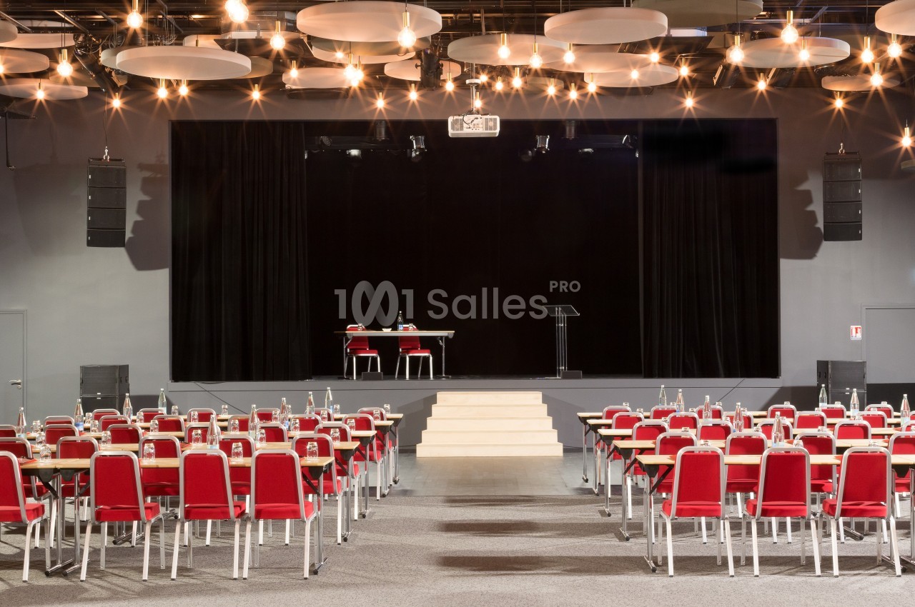 Salle de conférence avec des rangées de chaises rouges, une scène équipée d'une table, de chaises et d'un pupitre.