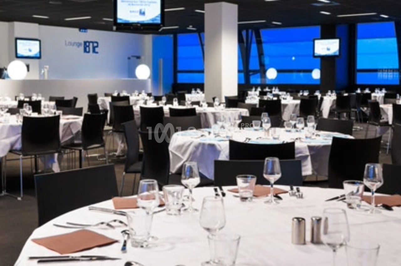 Salle de réception moderne avec tables rondes dressées, chaises noires et écrans accrochés aux murs.
