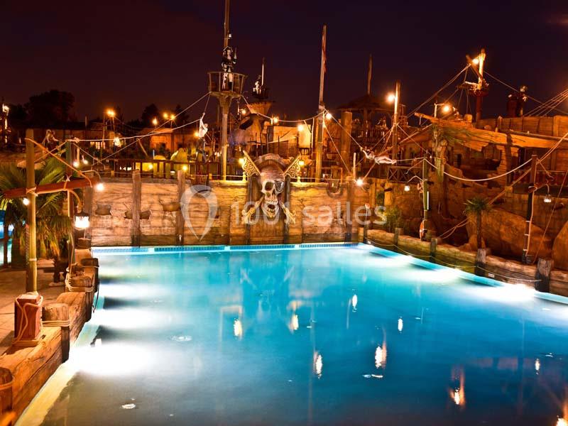 Piscine éclairée de nuit avec décorations de bateau pirate, cordages et crâne illuminé en arrière-plan.