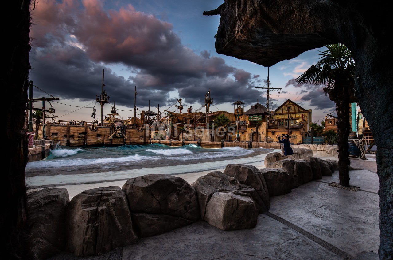 Vue d'un décor de port pirate avec des bateaux, des bâtiments en bois et des rochers sous un ciel nuageux.