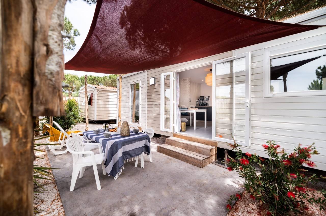 Terrasse d'un mobil-home avec table dressée, chaises blanches, voile d'ombrage rouge et végétation environnante.