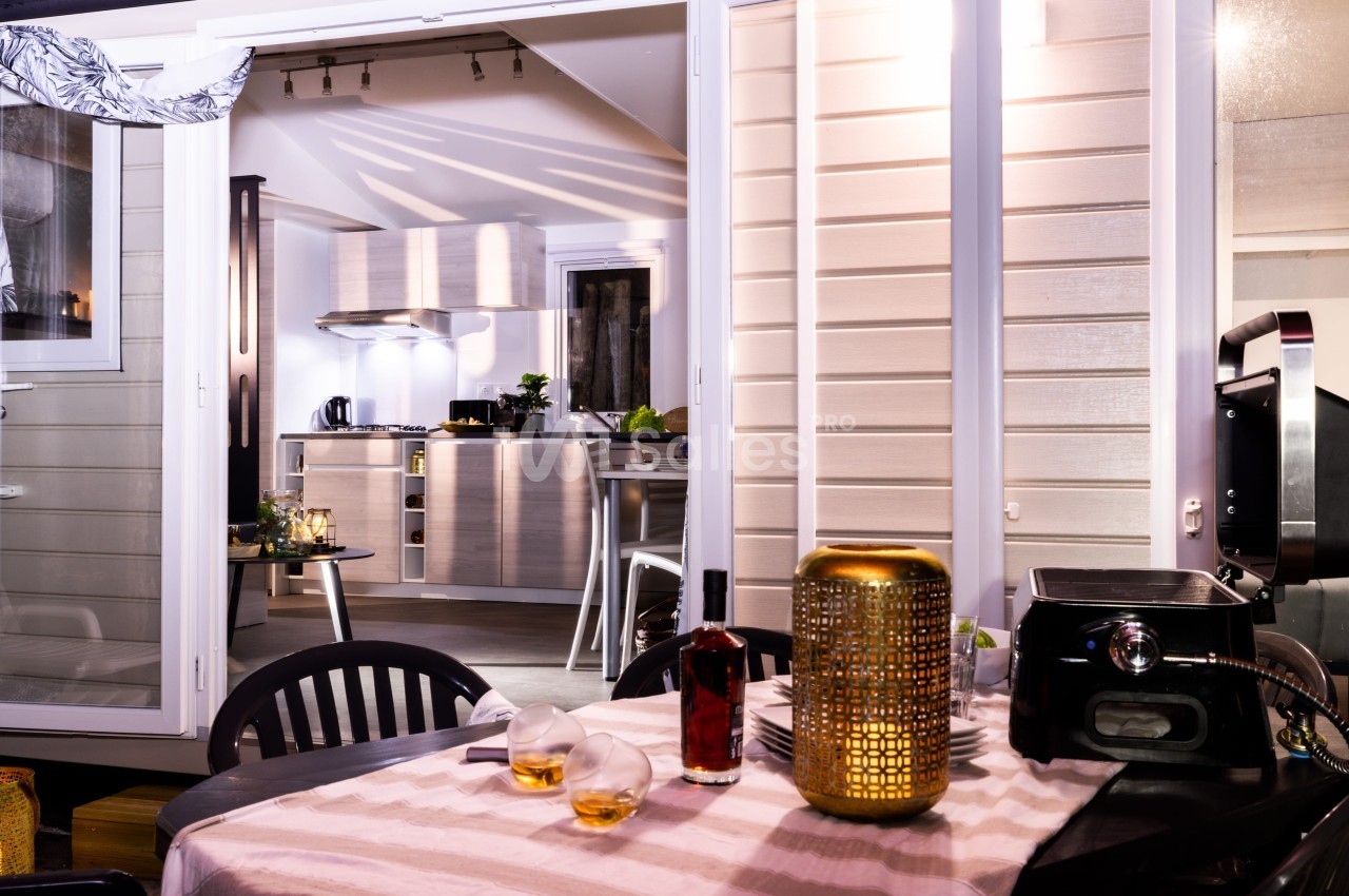 Vue d'une terrasse aménagée avec table dressée, donnant sur une cuisine moderne éclairée à l'intérieur.