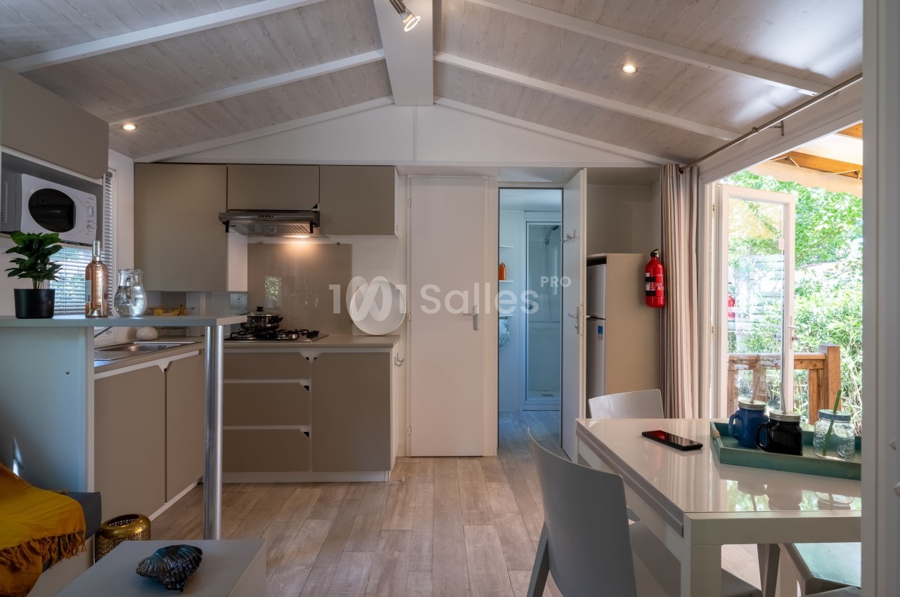Intérieur lumineux d'un mobil-home avec cuisine équipée, table à manger et porte ouverte sur une terrasse en bois.