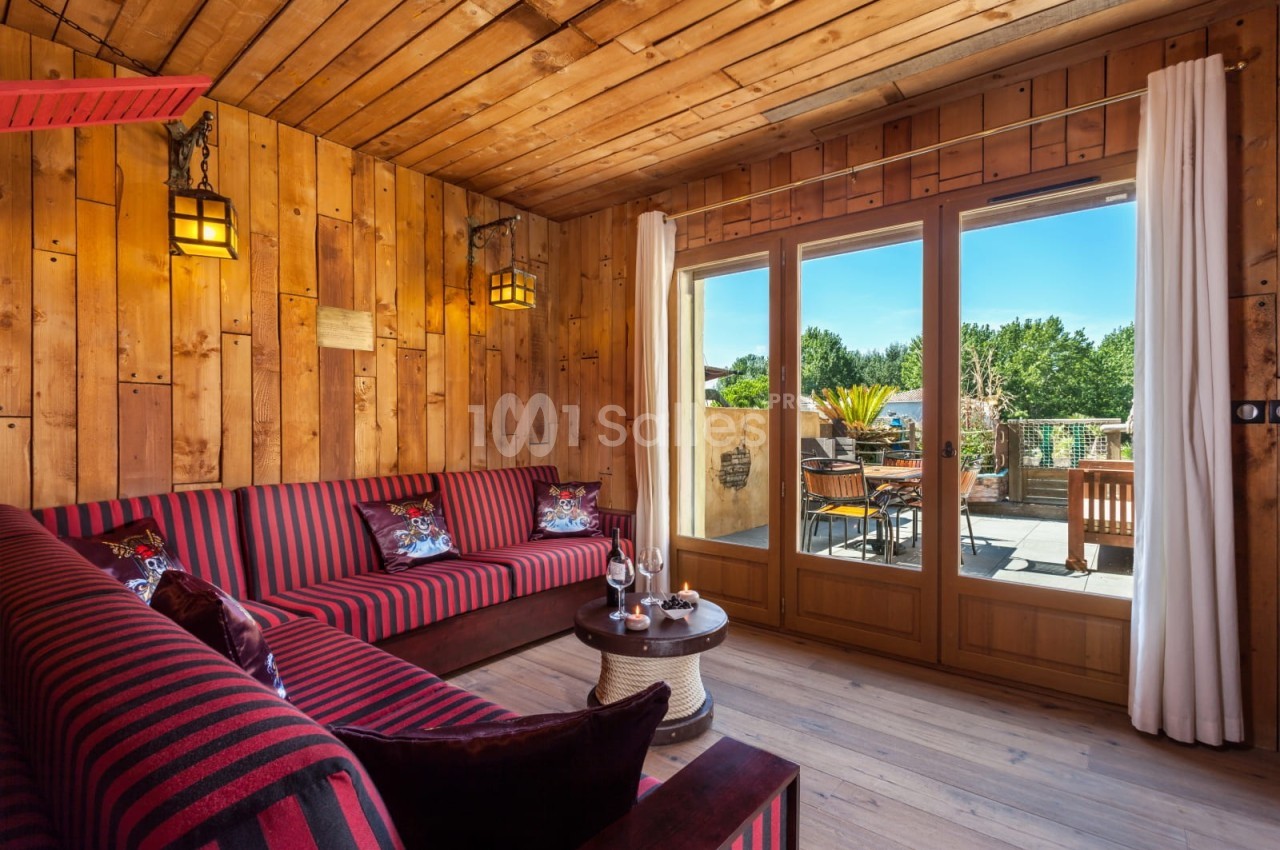 Salon avec murs en bois, canapé rayé rouge et noir, table basse, et grande baie vitrée donnant sur une terrasse ensoleillée.