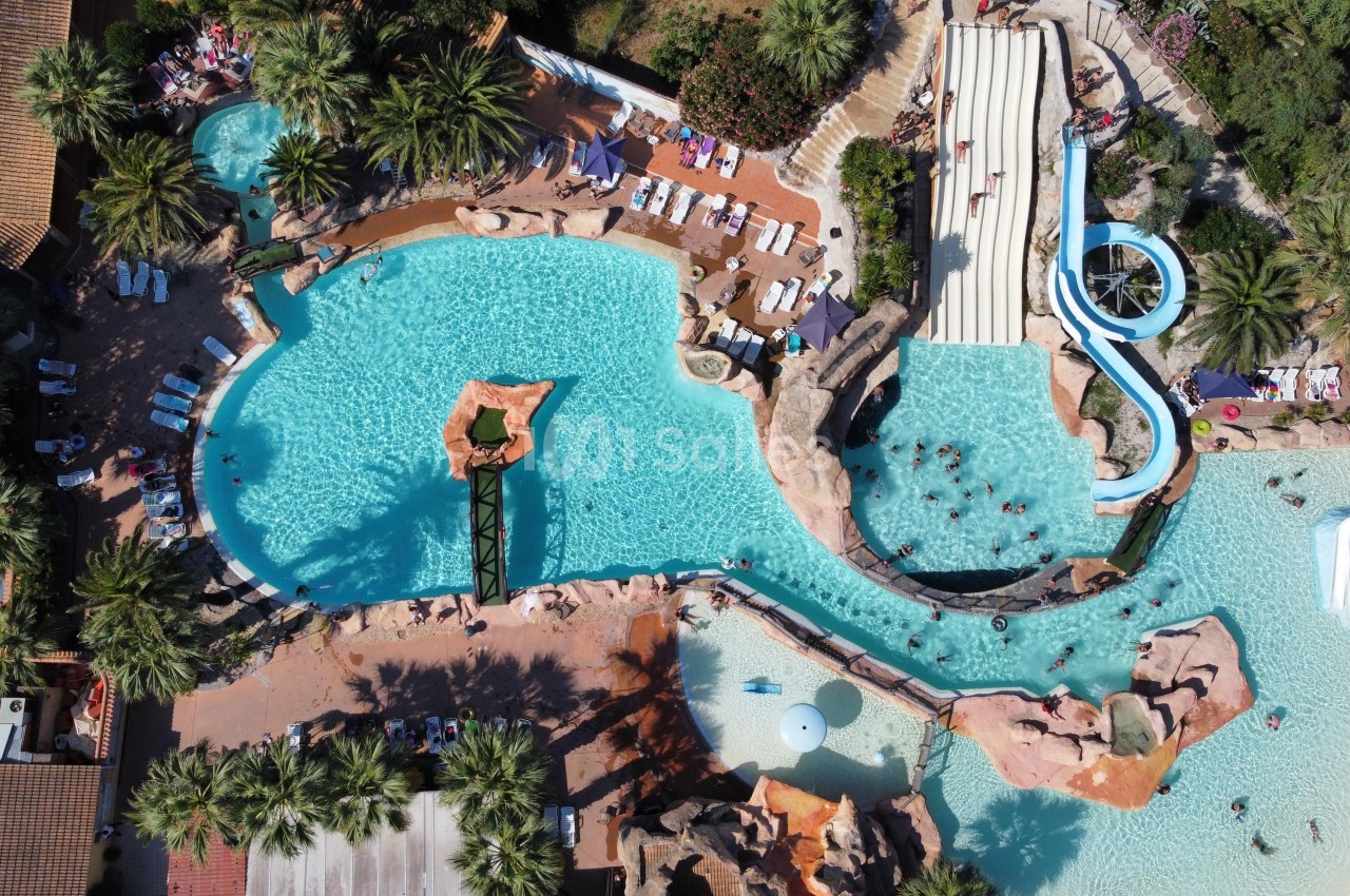 Vue aérienne d'un parc aquatique avec piscines, toboggans, rochers décoratifs et transats entourés de végétation.