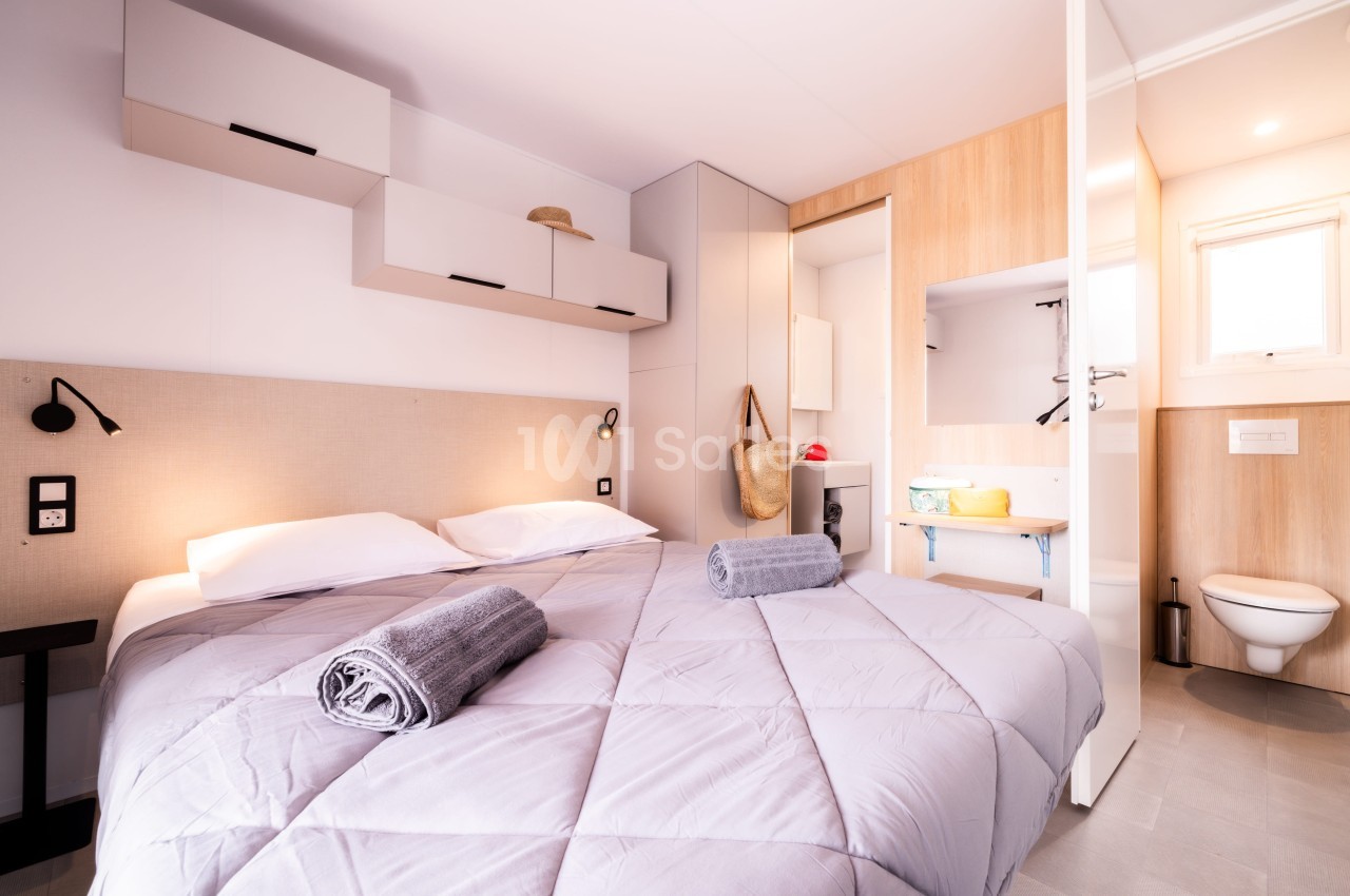 Chambre lumineuse avec lit double, draps gris, serviettes pliées, rangements muraux, et salle de bain attenante.