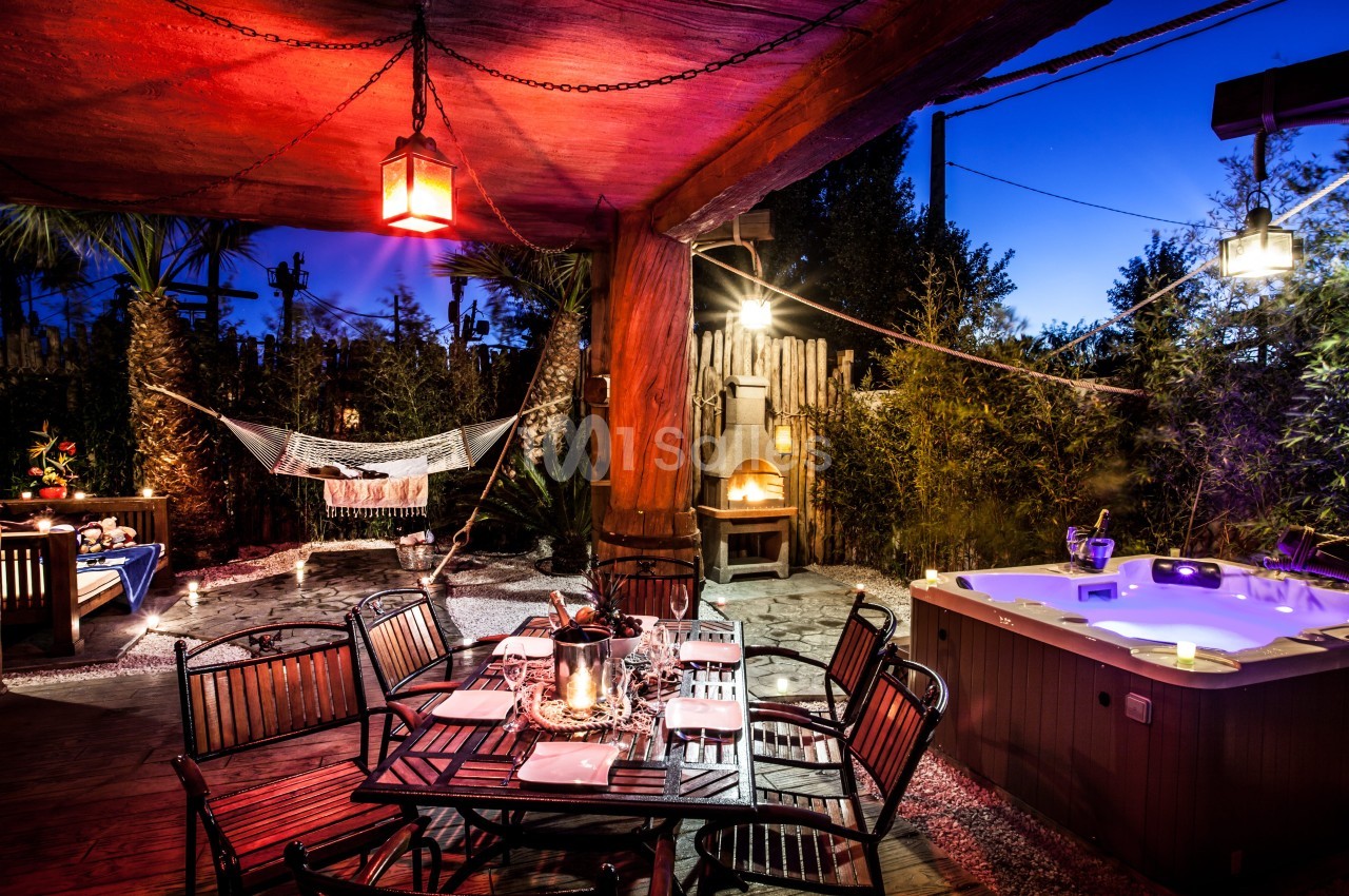 Terrasse éclairée de nuit avec table dressée, jacuzzi allumé, hamac suspendu et four extérieur en arrière-plan.