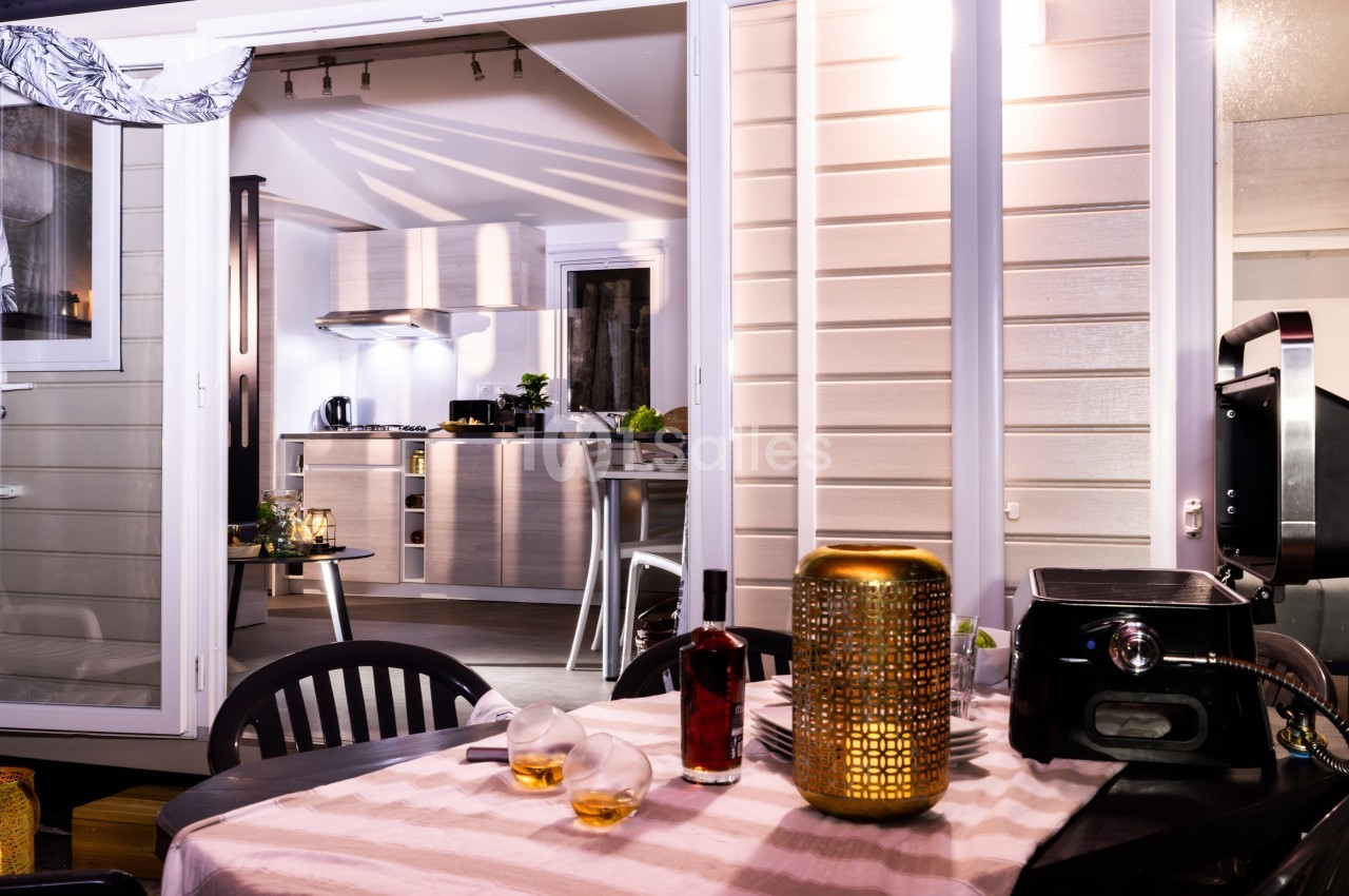 Vue d'une terrasse aménagée avec table dressée, donnant sur une cuisine moderne éclairée à l'intérieur.