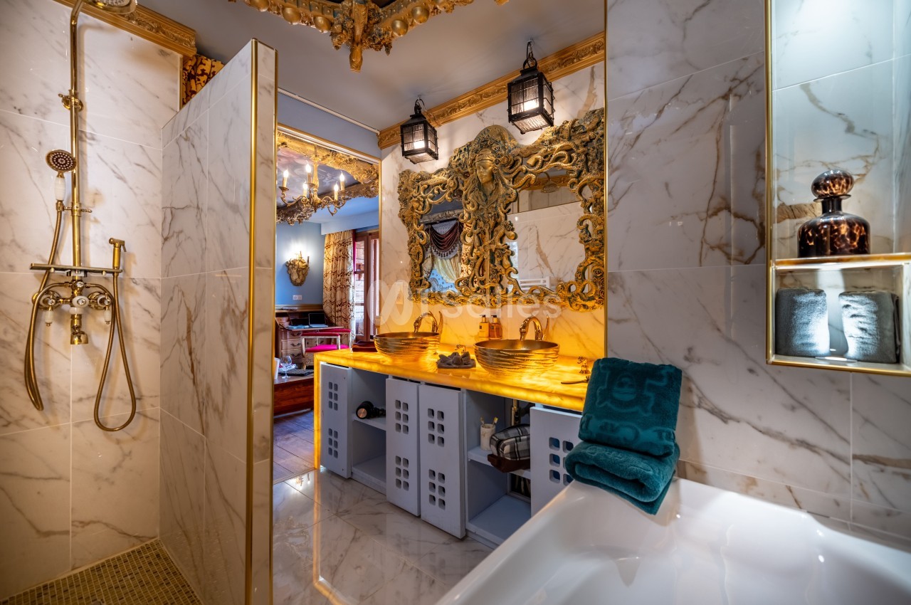 Salle de bain luxueuse avec murs en marbre, lavabo double doré, miroir orné et douche à robinetterie vintage.