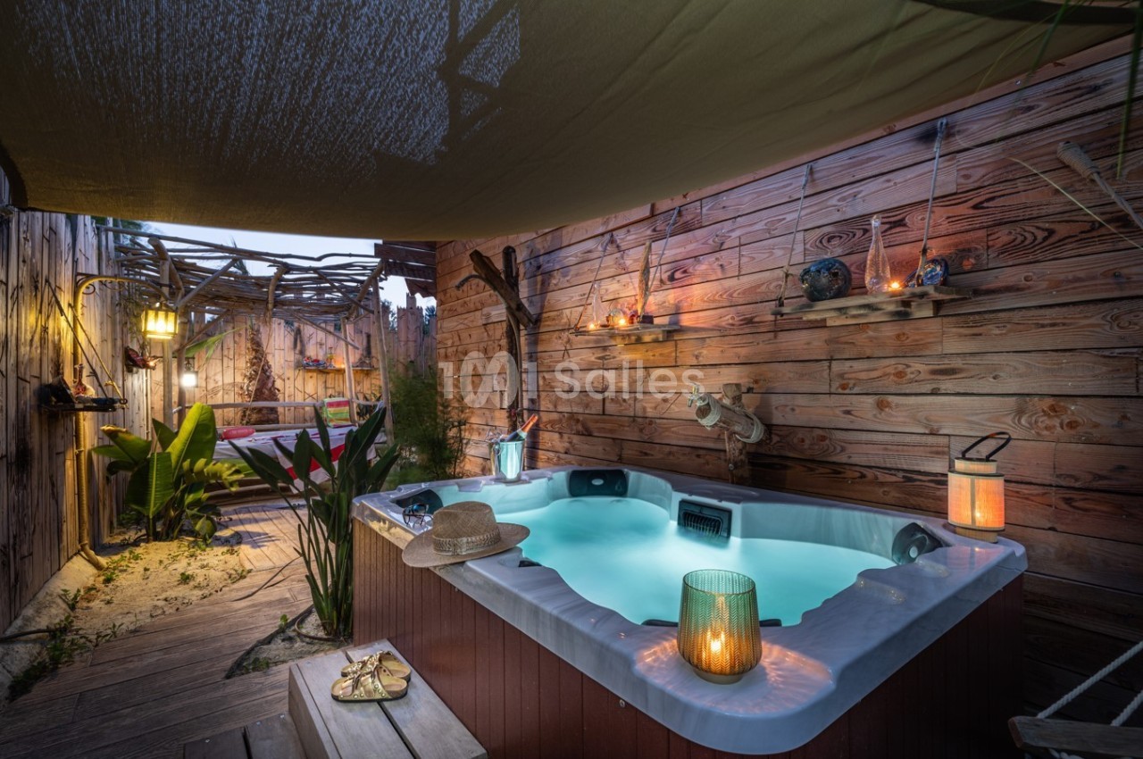 Jacuzzi extérieur éclairé le soir, entouré de bois et de plantes, avec des lanternes et une ambiance chaleureuse.