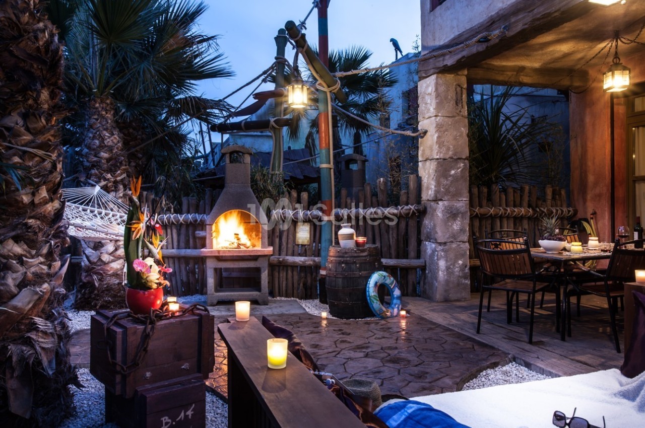 Terrasse extérieure avec cheminée allumée, table dressée, plantes tropicales et éclairage tamisé en soirée.
