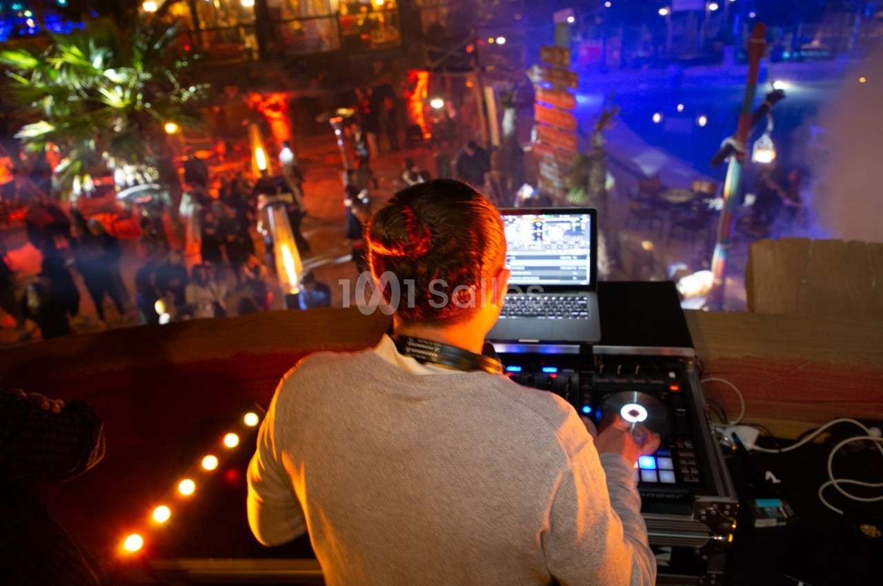Un DJ mixe de la musique sur une console, face à une foule dans un lieu animé et éclairé par des lumières colorées.