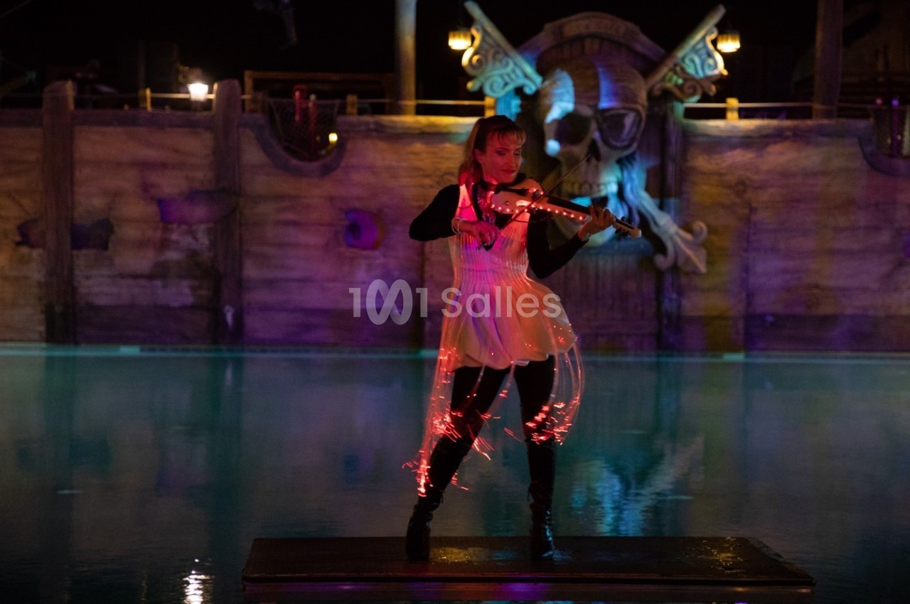 Une violoniste joue sur une scène flottante éclairée, devant un décor de bateau pirate illuminé.