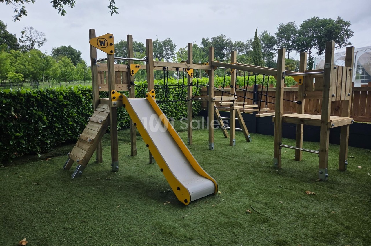 Structure de jeu en bois avec toboggan, pont suspendu et filets, installée sur une pelouse artificielle entourée de haies.