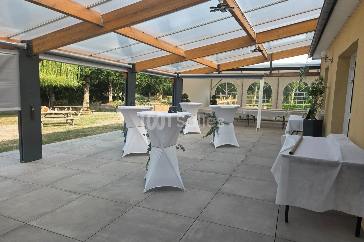 Terrasse couverte avec tables hautes drapées de blanc, entourée de verdure et éclairée par une lumière naturelle.