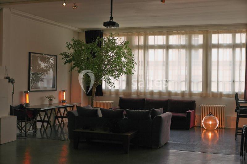 Espace de vie lumineux avec canapé, table, plantes et grandes fenêtres laissant entrer la lumière naturelle.