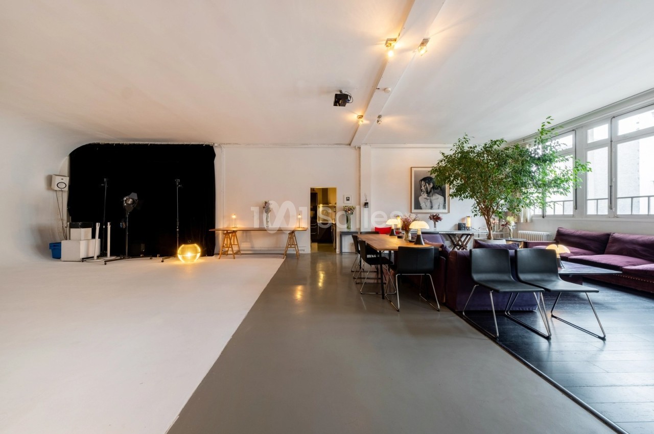 Espace intérieur lumineux avec coin salon, grande table, plantes et zone de studio photo équipée de projecteurs.