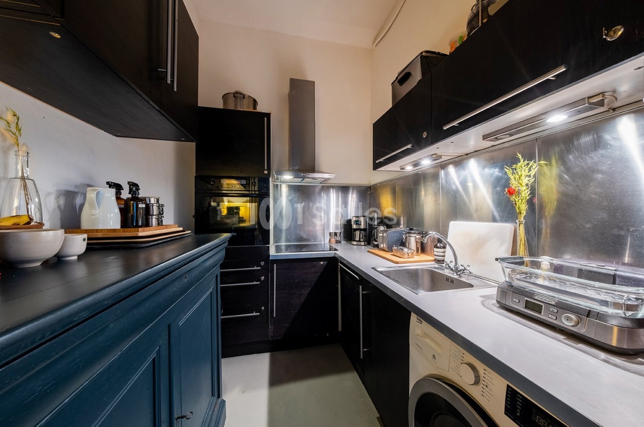 Cuisine moderne avec meubles noirs, plan de travail en bois, électroménagers et crédence métallique réfléchissante.