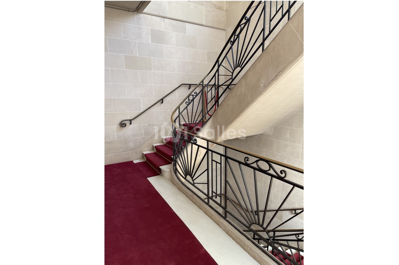 Escalier en pierre avec tapis rouge et rampe en fer forgé, dans un intérieur lumineux aux murs clairs.
