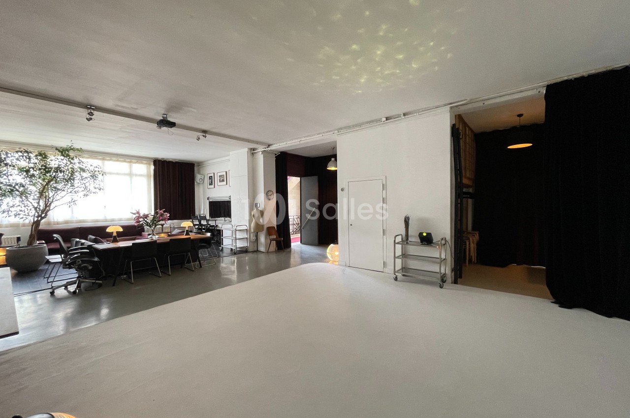 Espace intérieur lumineux avec un grand salon, une table à manger, des plantes et des rideaux noirs en arrière-plan.