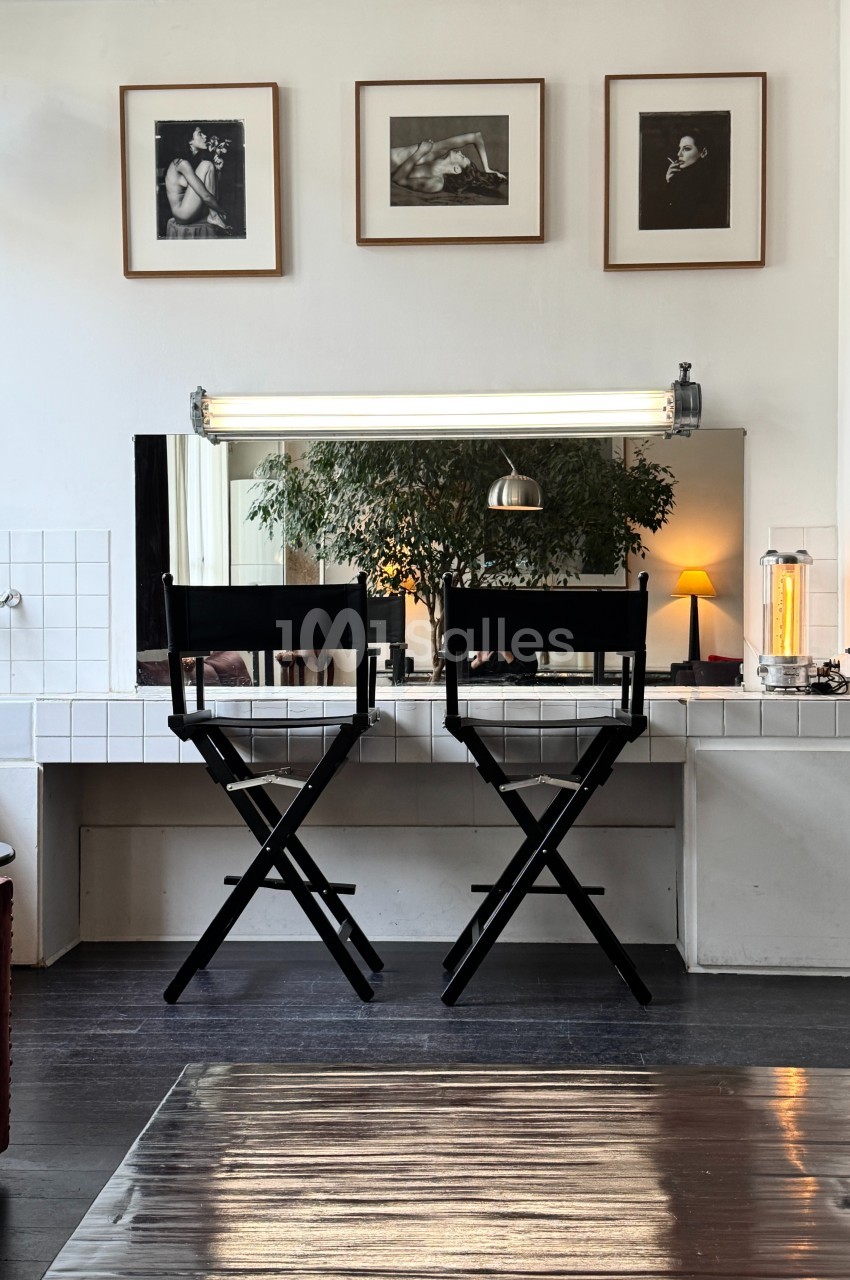 Deux chaises hautes noires devant un miroir éclairé, avec des photos encadrées accrochées au mur blanc.