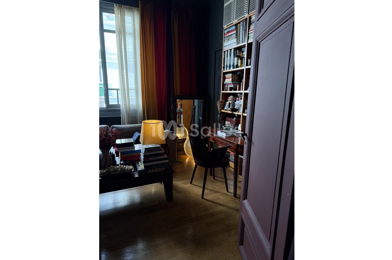 Pièce sombre avec bibliothèque, bureau en bois, lampe allumée et fauteuil, près d'une fenêtre avec rideaux.
