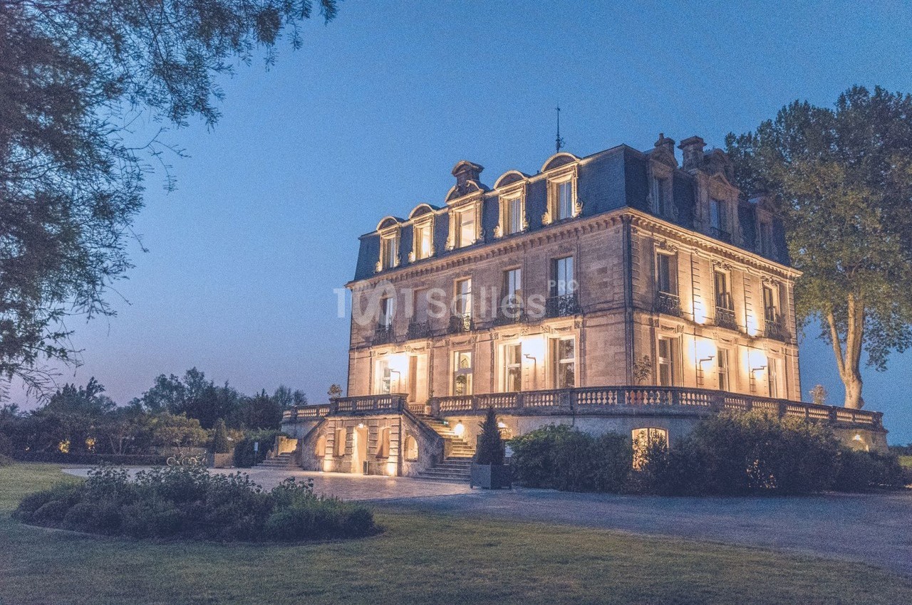 Manoir éclairé au crépuscule, entouré de verdure et d'arbres, avec un ciel bleu en arrière-plan.