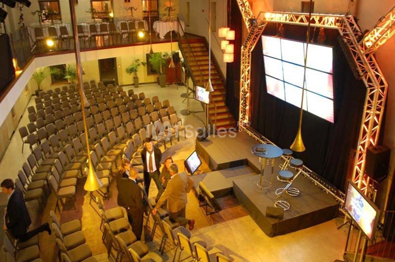 Salle de conférence avec sièges alignés, scène équipée d'écrans et de projecteurs, quelques personnes discutant.