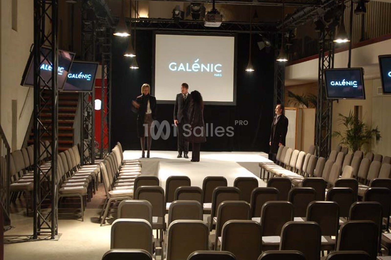 Salle de conférence avec une scène centrale, des sièges alignés et des écrans affichant ’Galénic Paris’.