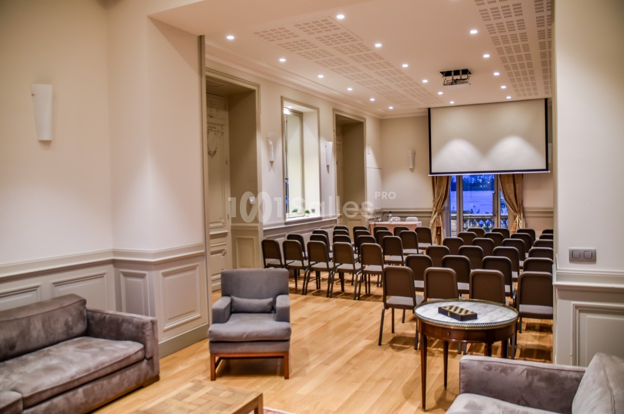 Salle de conférence lumineuse avec rangées de chaises, écran de projection et coin salon aménagé.