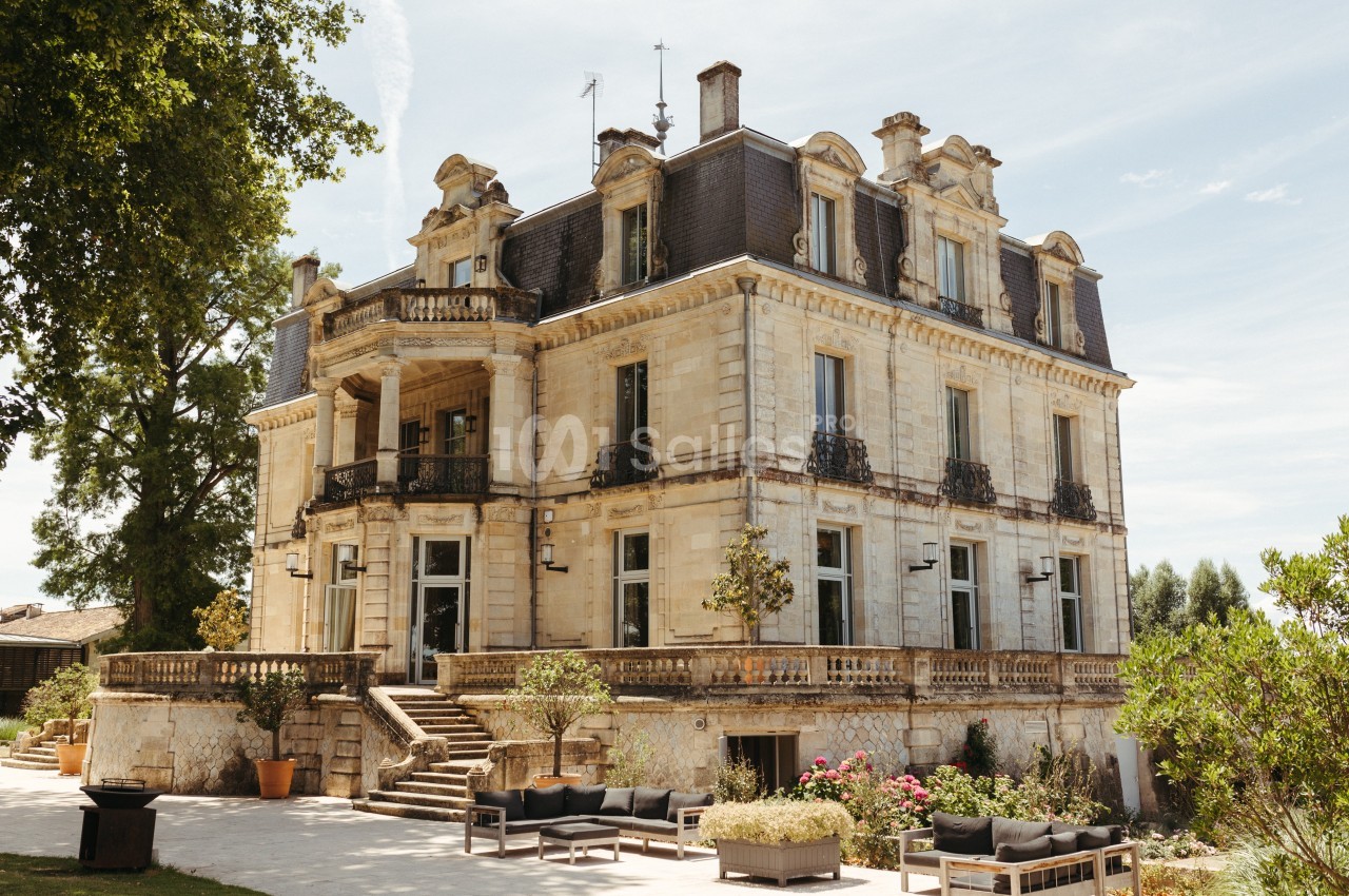 Façade d'un manoir en pierre de style classique avec balcons, entouré de végétation et mobilier d'extérieur.