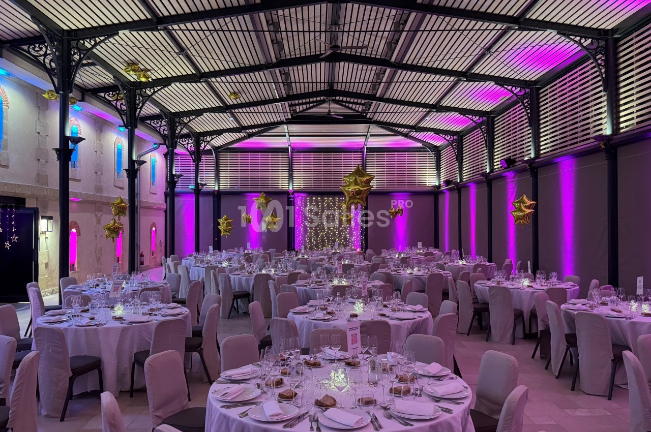 Salle de réception décorée avec des tables rondes dressées, éclairage violet et doré, et ballons en forme d'étoiles.
