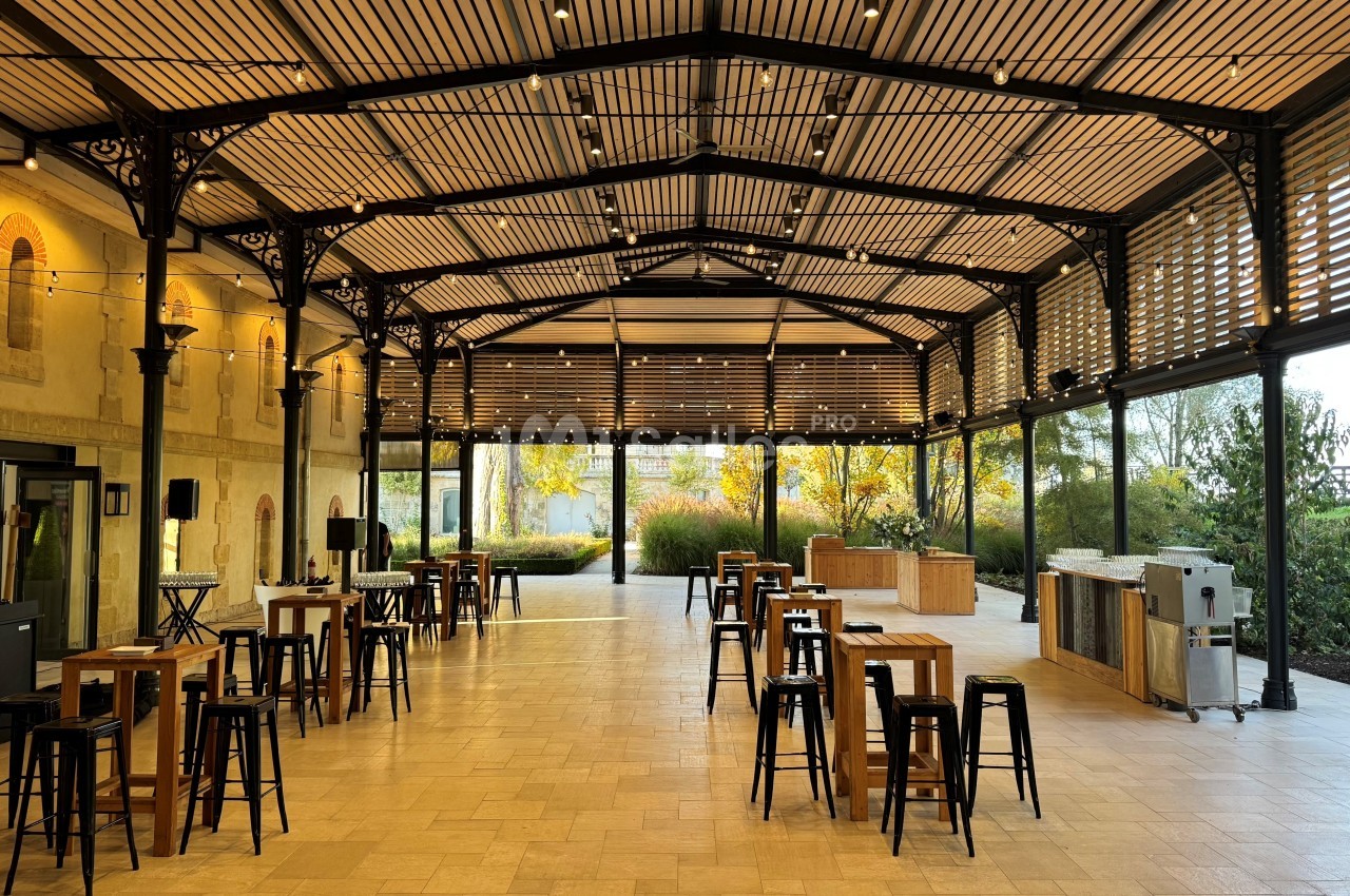 Salle lumineuse avec toit en verre, tables hautes en bois et tabourets noirs, entourée de verdure extérieure.