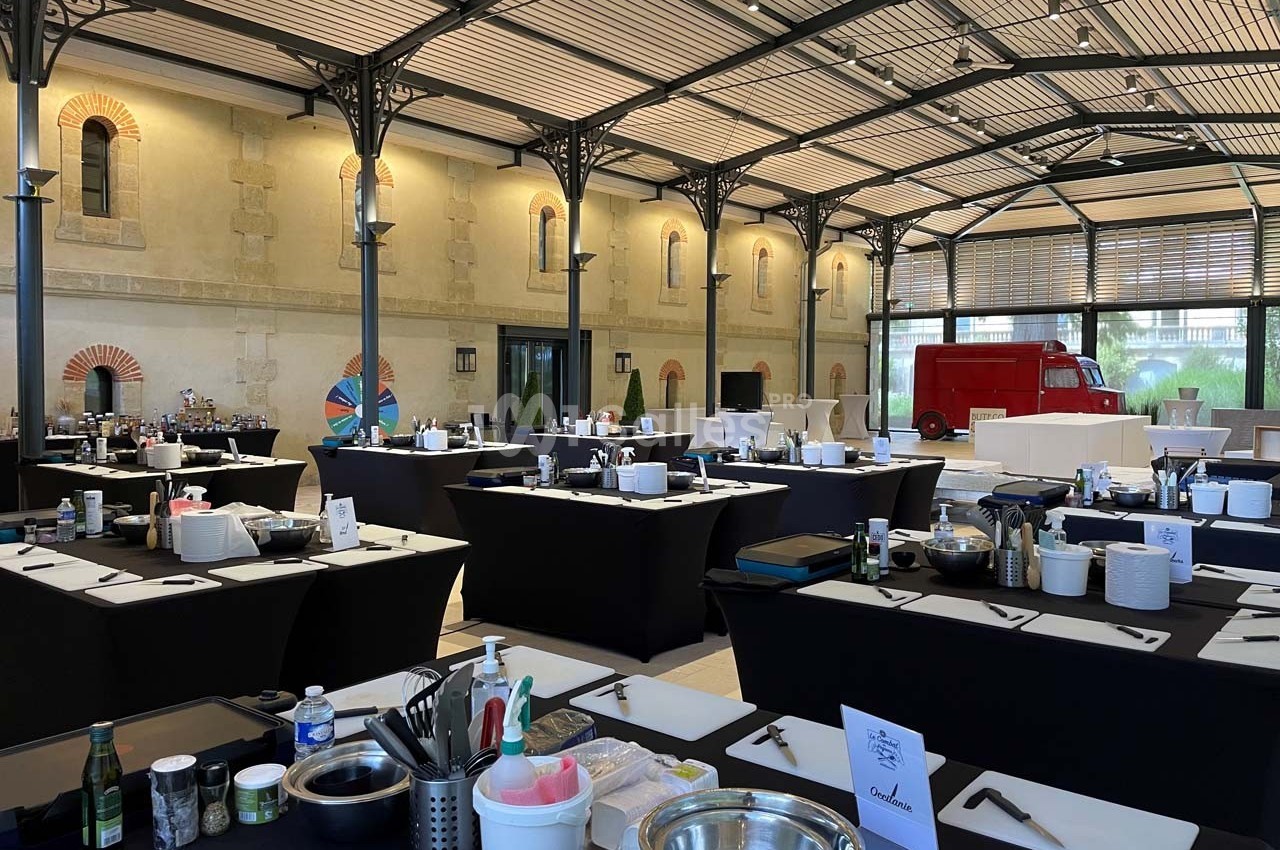 Salle aménagée avec des tables noires équipées de matériel de cuisine, sous une verrière avec un camion rouge en arrière…