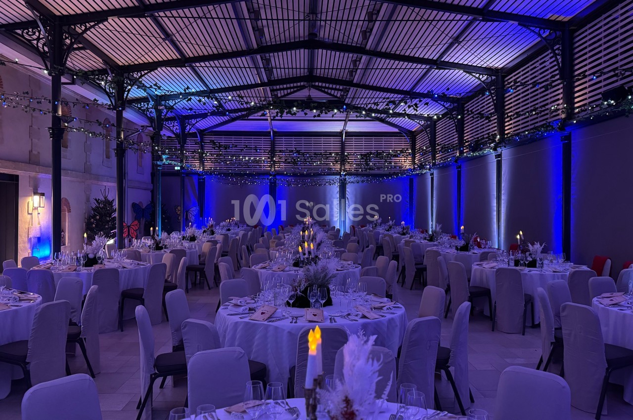 Salle de réception élégamment décorée avec des tables rondes dressées, éclairage bleu et guirlandes lumineuses suspendues.