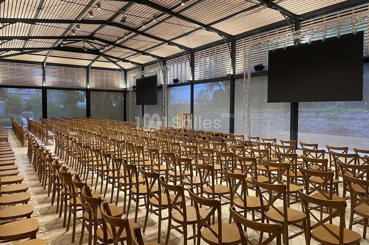 Salle de conférence vide avec des rangées de chaises en bois, un pupitre central et deux écrans noirs suspendus.