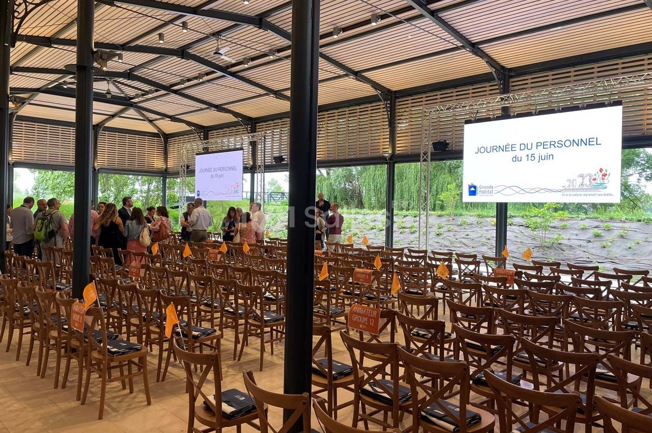 Salle avec des chaises en bois, écrans affichant ’Journée du personnel du 15 juin’ et participants debout au fond.