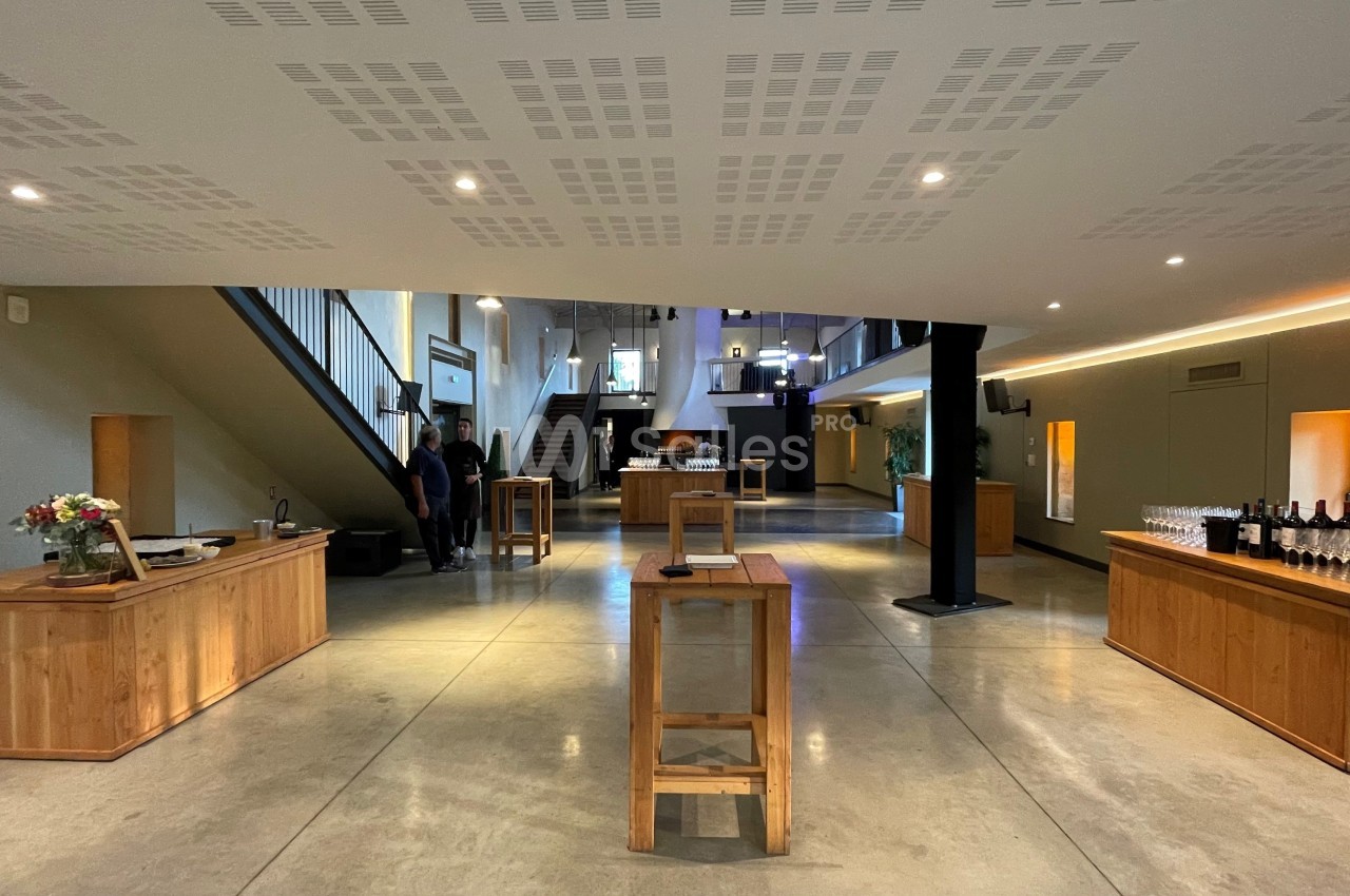 Salle lumineuse avec mobilier en bois, tables hautes, escalier et espace aménagé pour une réception ou un événement.