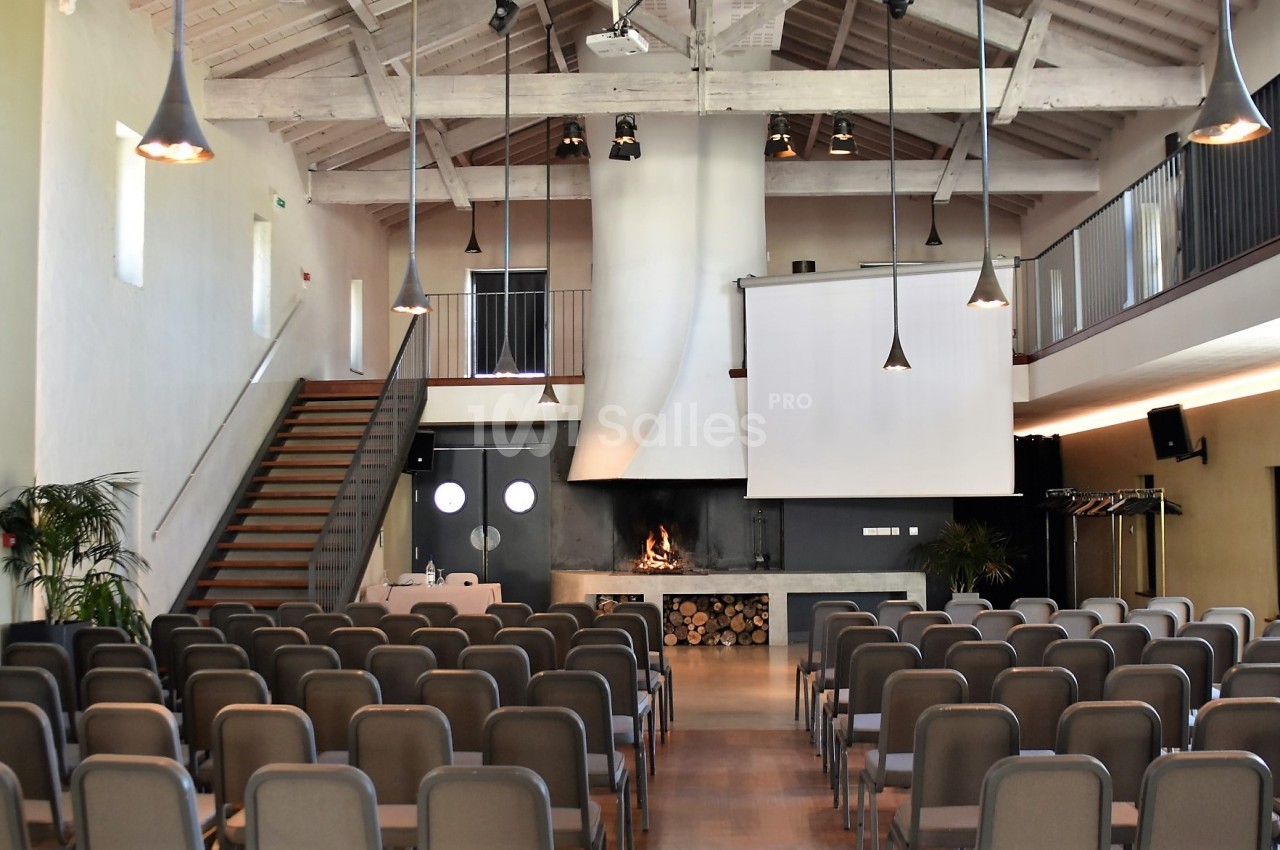 Salle de conférence lumineuse avec rangées de chaises, écran de projection, cheminée et mezzanine.