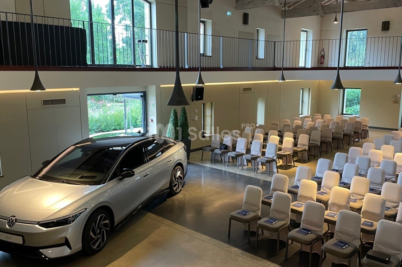 Voiture argentée exposée dans une salle lumineuse avec des rangées de chaises disposées pour un événement.