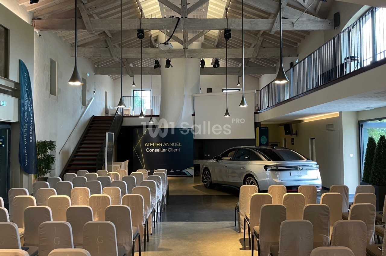 Salle de conférence avec des rangées de chaises, une scène, un écran de projection et une voiture exposée.