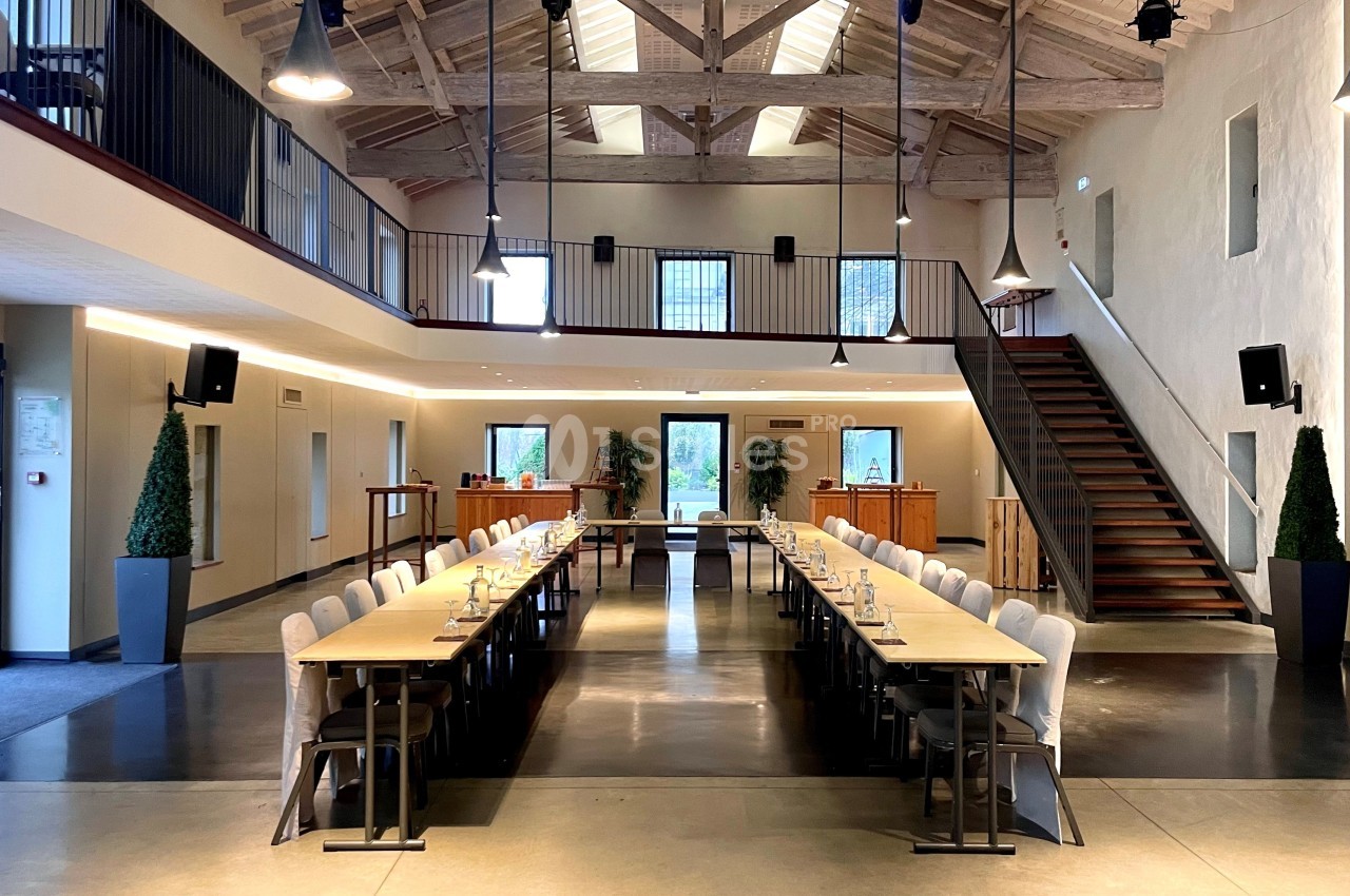 Grande salle lumineuse avec tables alignées en U, chaises, escalier en bois et plafond avec poutres apparentes.