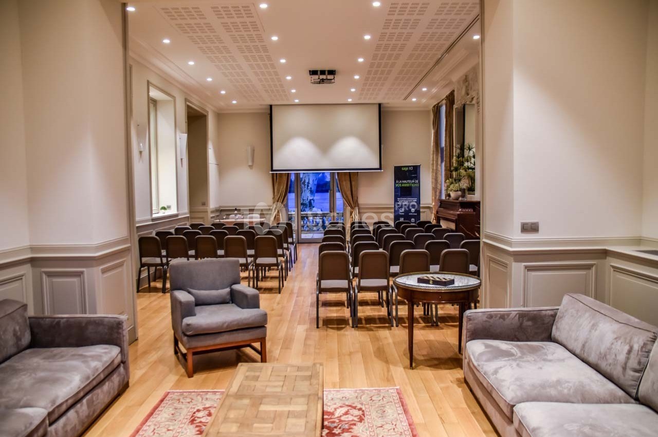 Salle de conférence lumineuse avec rangées de chaises, écran de projection et coin salon en premier plan.
