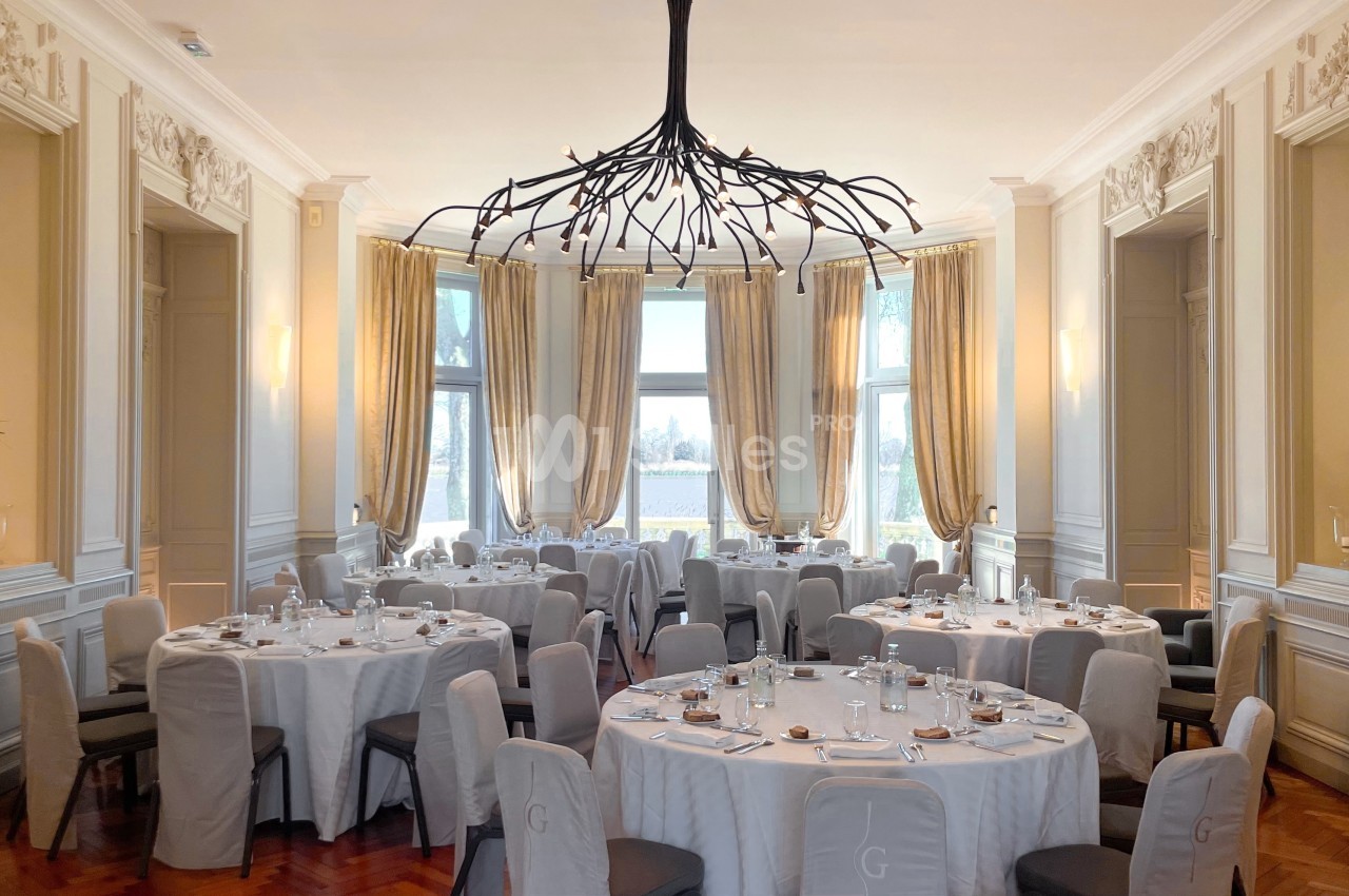 Salle élégante avec tables rondes dressées, chaises couvertes, grandes fenêtres et lustre moderne en forme de branches.