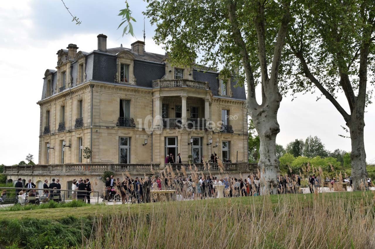 Façade d'un château en pierre entouré d'arbres, avec des personnes rassemblées sur l'esplanade devant.