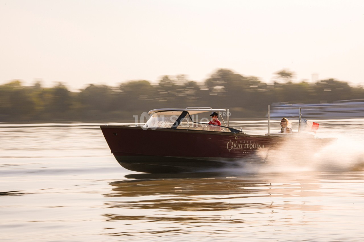 Un bateau à moteur rouge navigue rapidement sur une rivière, avec deux personnes à bord et des arbres en arrière-plan.
