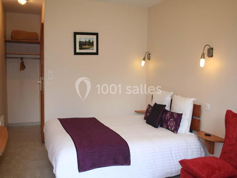 Chambre avec lit double, couvre-lit violet, coussins décoratifs, éclairage mural et placard ouvert en arrière-plan.