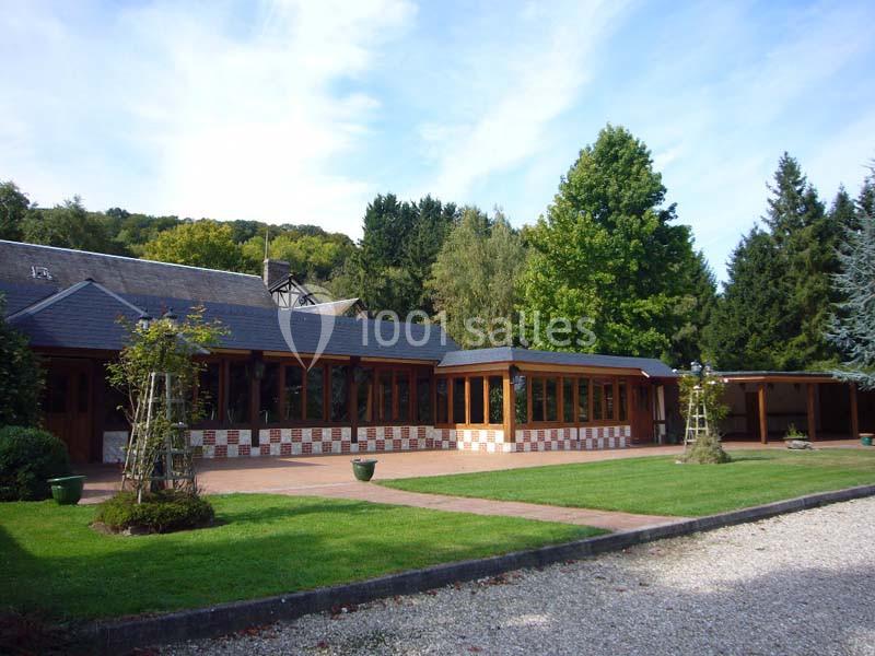 Grande maison avec façade en bois, terrasse pavée, pelouse et arbres environnants sous un ciel dégagé.