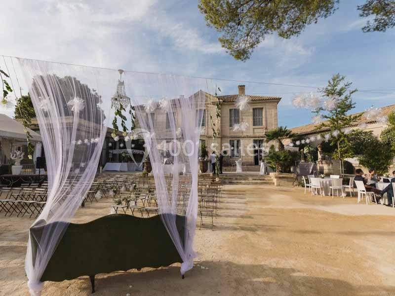 Cour extérieure aménagée pour un événement avec chaises, tables, voilages blancs et bâtiment en arrière-plan.