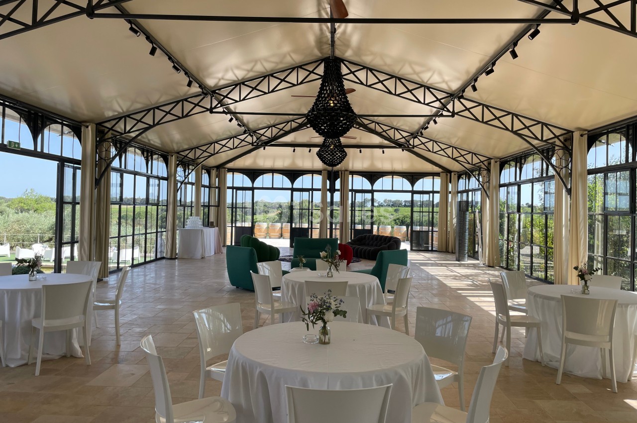 Salle lumineuse avec toit en toile, grandes baies vitrées, tables rondes dressées et canapés en fond.